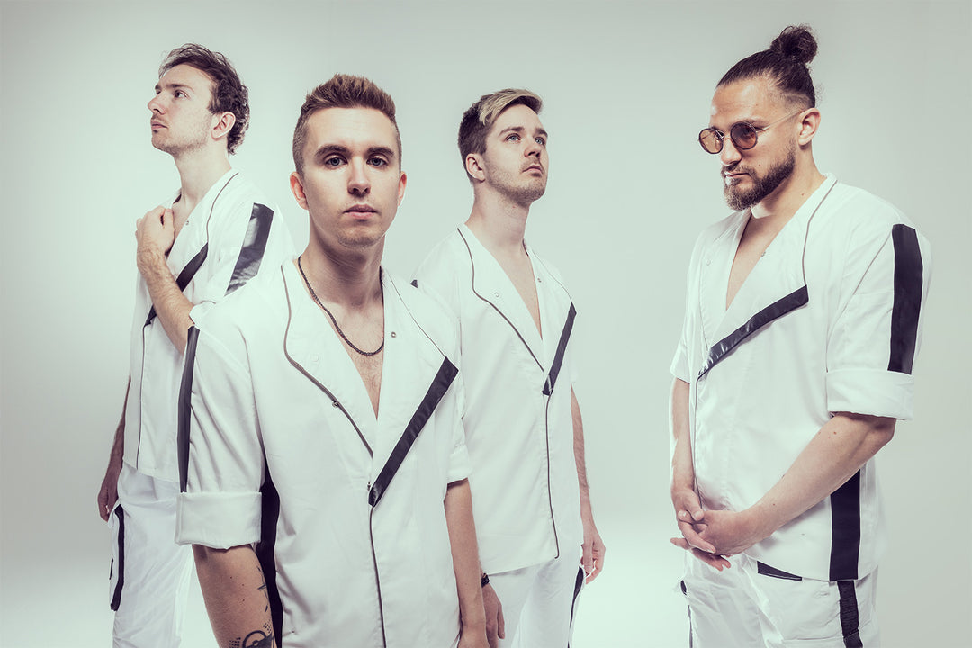 Four men wearing matching white outfits with black accents stand against a plain light background. Three look in different directions; one wears glasses and has his hair in a bun.