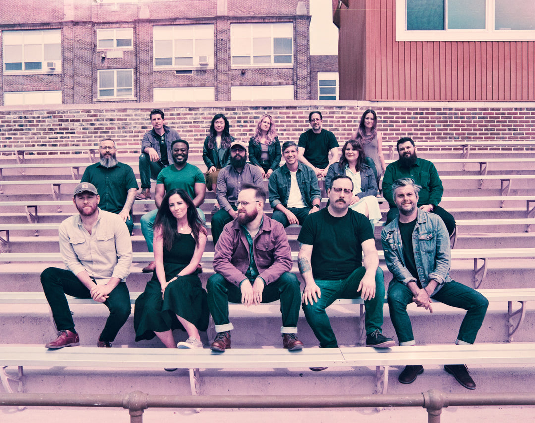 A group of seventeen adults sits on bleachers outdoors, posing together and smiling for a group photo, with a brick building in the background.