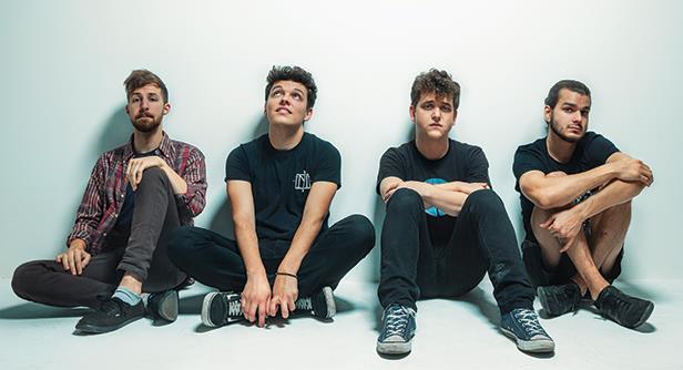 Four young men sit on the floor against a plain white wall, wearing casual clothes and sneakers, looking relaxed and thoughtful.
