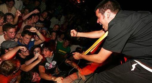 A musician passionately plays guitar on stage while an energetic crowd cheers and raises their fists at a lively concert.