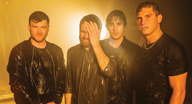 Four men stand together in the rain, wearing dark wet clothes, illuminated by a warm yellow light in the background. One man wipes his face while the others look toward the camera.