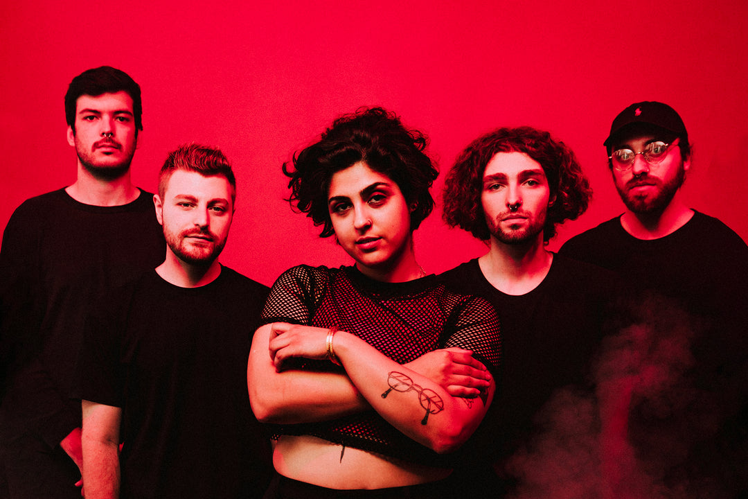 Five people in black clothing stand against a vibrant red background. The person in front crosses their arms, and the group looks serious. There is a light haze or smoke at the bottom right.