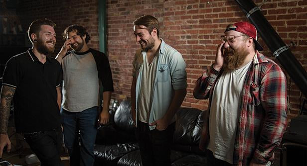 Four men stand together indoors, laughing and smiling near a black couch. The setting features exposed brick walls, and they appear relaxed and friendly, enjoying each others company.