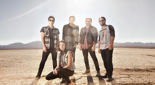 Five people pose together in a dry, cracked desert landscape under a bright sun. Four stand while one crouches in front, all wearing casual, modern clothing with mountains in the background.