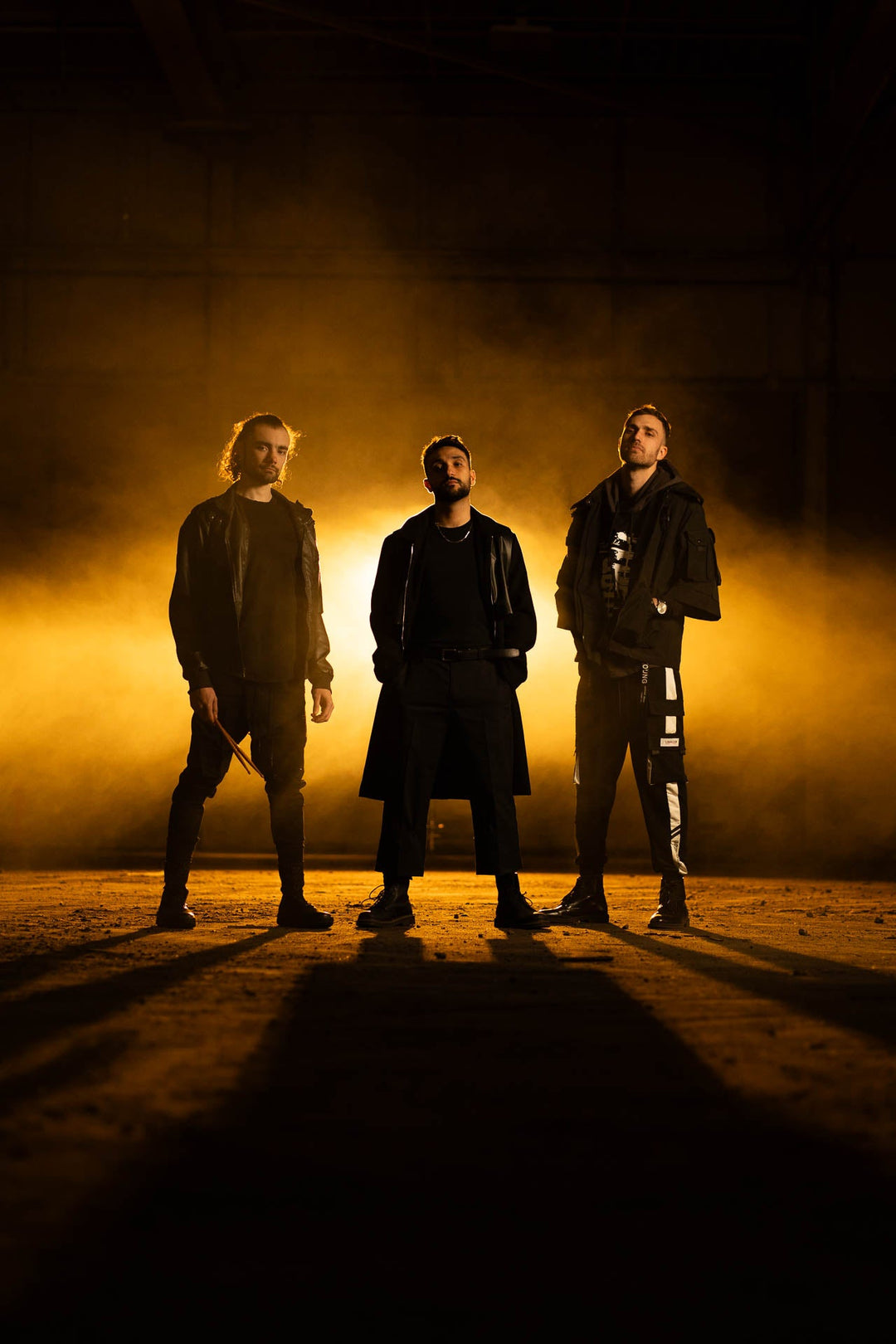 Three men stand confidently in a dramatic setting, illuminated by a backdrop of yellow-orange fog and low lighting, casting strong shadows and creating a moody, cinematic atmosphere.