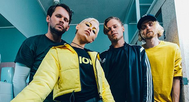 Four people stand close together in a brightly-lit room, facing the camera. One wears a yellow jacket and leans forward, while the others wear casual shirts and jackets, with serious expressions.
