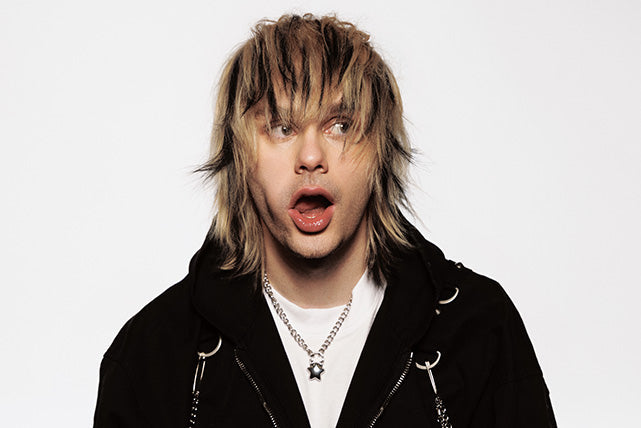 A person with shaggy, two-toned blond and brown hair wearing a black hoodie and silver chain necklace looks surprised with their mouth open against a plain white background.