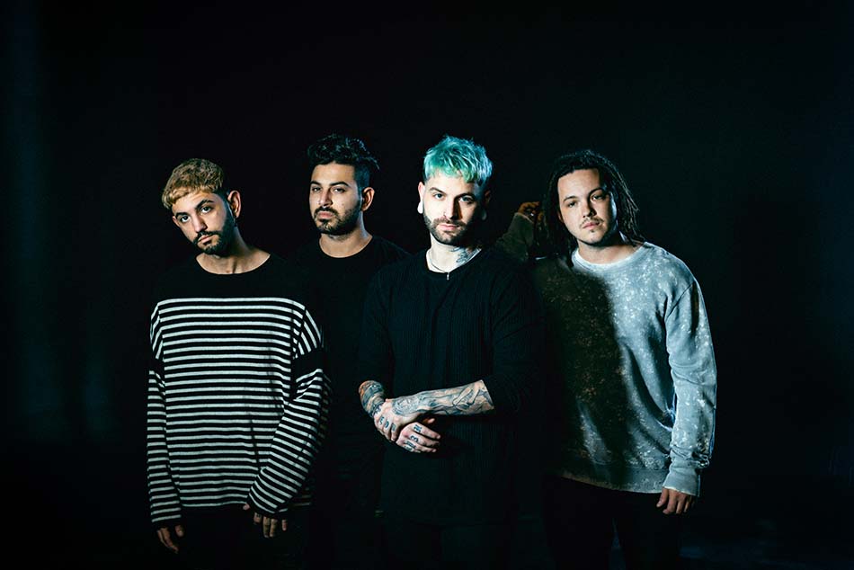 Four men stand together against a dark background; one has blue hair, another wears a striped sweater, and the group appears serious, lit dramatically from the front.