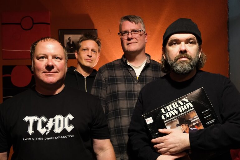 Four men pose indoors in front of Rock Against Bush posters; one man kneels while the others stand. One wears an Against Me! shirt, lifting it to reveal his stomach and holding a drink.