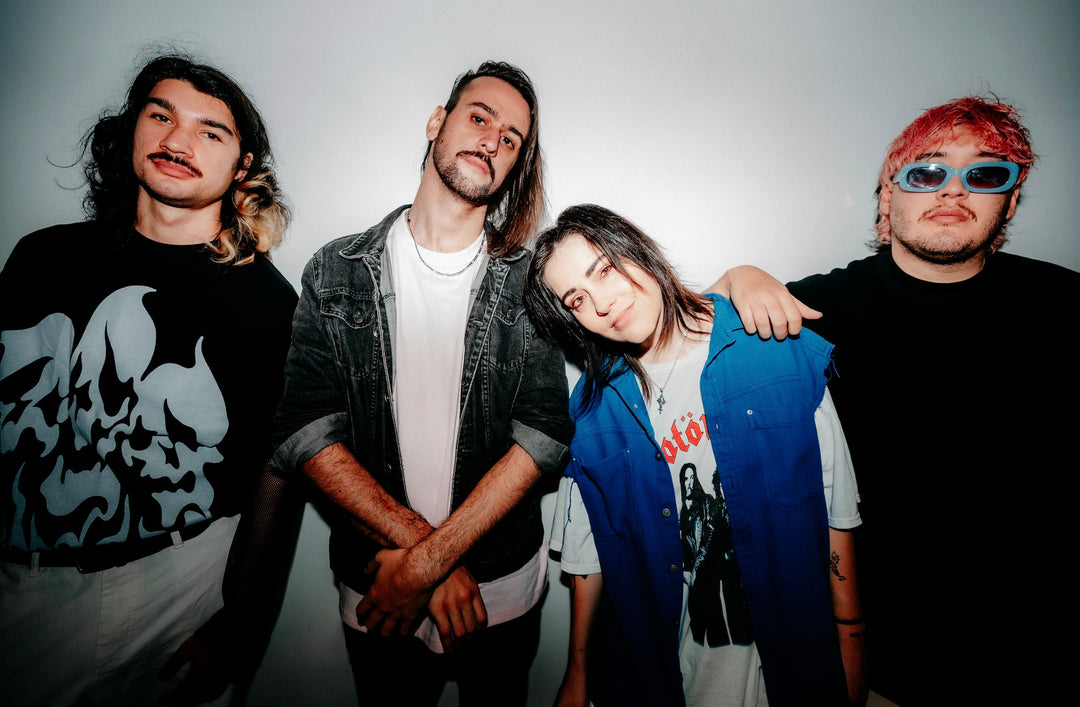 Four young adults pose against a white background, standing closely together. One person has their arm around anothers shoulder. They wear casual clothes and have various hairstyles and expressions.