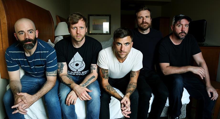 Five men sit closely together on a bed in a dimly lit room, looking at the camera. They wear casual shirts and jeans, and some have beards or tattoos. A lamp and mirror are visible in the background.