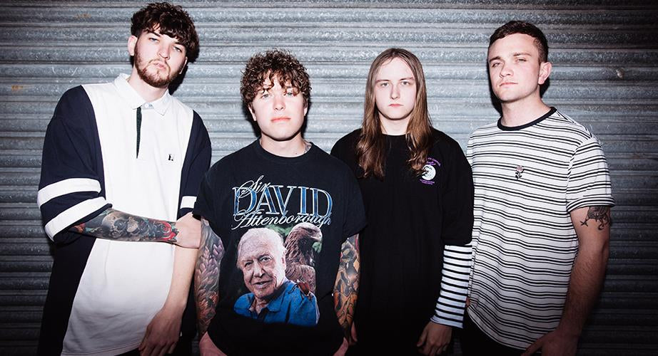 Four young men stand in front of a metal roller door, wearing casual clothes; one wears a David Attenborough shirt, and two have visible tattoos. They look directly at the camera with neutral expressions.