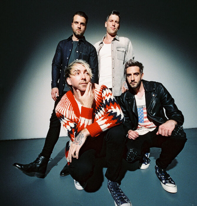 Four men in dark jackets stand in front of a wall covered with black graffiti. One man holds a spray paint can toward the camera, and the group poses confidently.