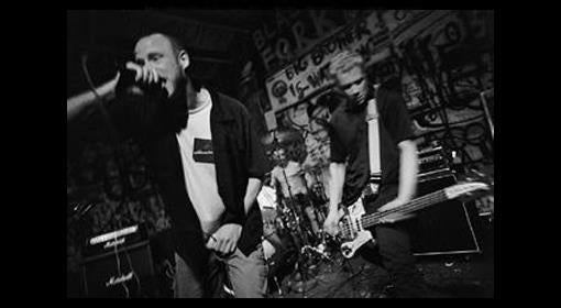A black and white photo of a punk band performing on stage; the vocalist sings into a microphone while the bassist plays energetically. Graffiti covers the wall behind the drum set.
