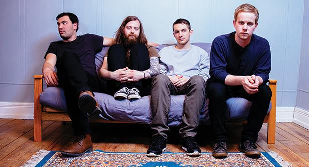 Four men sit on a blue couch against a light blue wall, looking in different directions. One has long hair and a beard, while the others have short hair. A patterned rug is on the wooden floor.