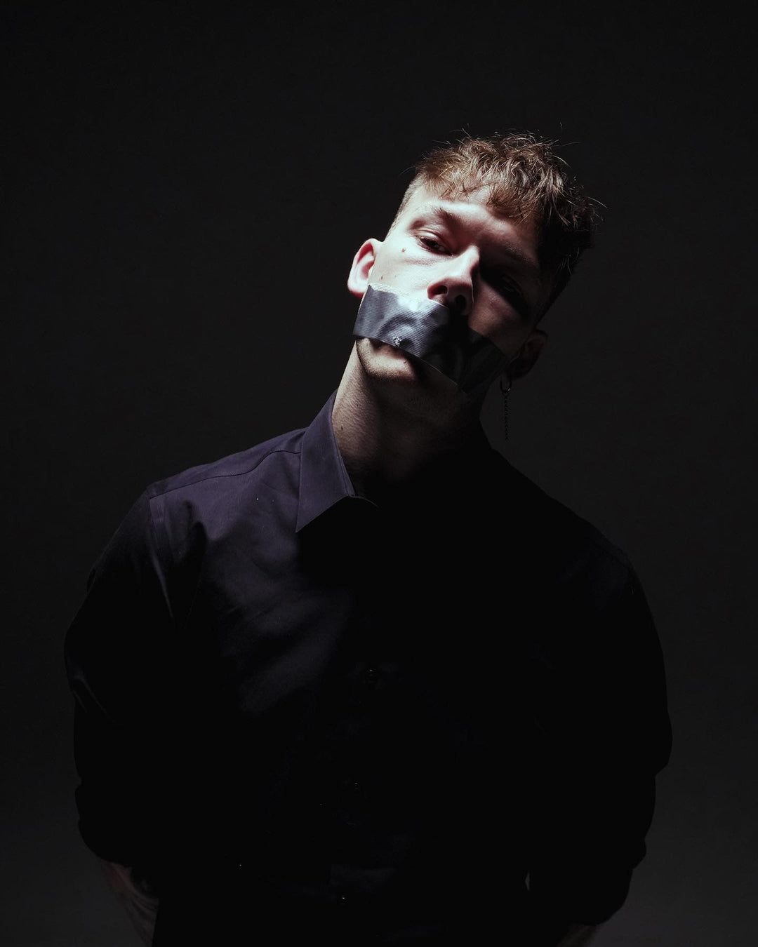 A person in a black shirt stands against a dark background with silver tape covering their mouth, head tilted slightly, and expression serious, creating a dramatic and intense mood.