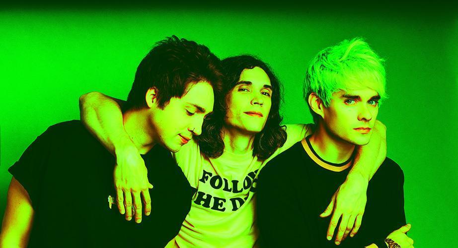 Three young men pose together in front of a green background, with the person in the middle resting his arms on the shoulders of the other two. The lighting gives the image a neon green tint.