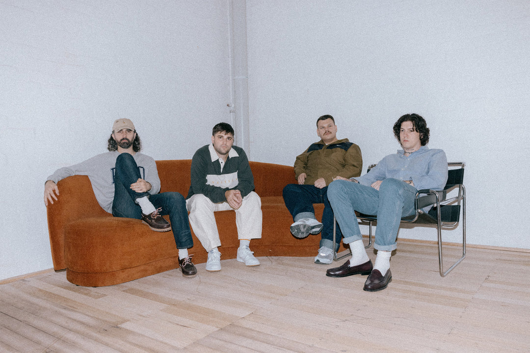 Four men sit relaxed on a brown couch and a black chair in a minimalist, white-walled room with wooden floors. They are casually dressed and looking toward the camera.