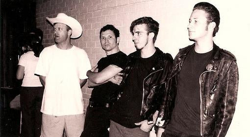 Four men stand against a brick wall; one wears a cowboy hat and white shirt, the others wear dark shirts and leather jackets, looking in different directions. The photo is in black and white.