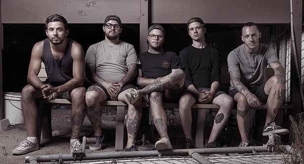 Five men with tattoos sit on benches outside, facing the camera. They are casually dressed in T-shirts, shorts, and caps, with neutral expressions and relaxed postures.