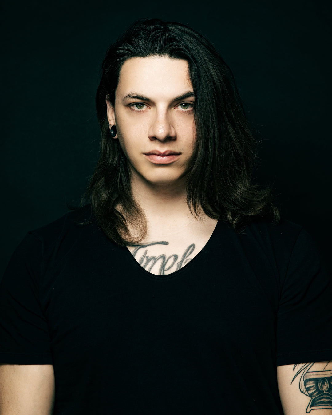 A person with long dark hair, light eyes, and ear gauges, wearing a black shirt, stands against a dark background. They have a tattoo across their chest, partly visible above the shirt collar.