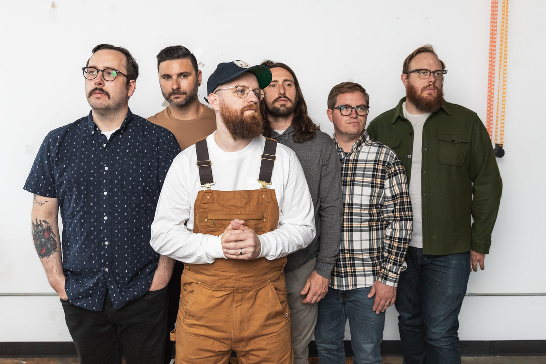 Six men stand side by side against a plain white wall, looking in different directions. They wear casual clothing, including patterned shirts, a cap, and overalls.