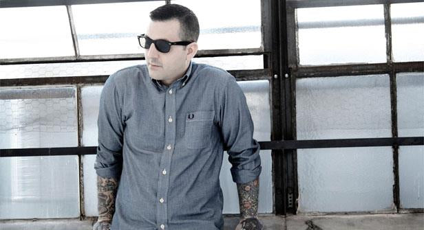 A person wearing sunglasses and a blue button-up shirt with rolled sleeves, showing tattooed arms, stands in front of large industrial-style windows, looking to the side.