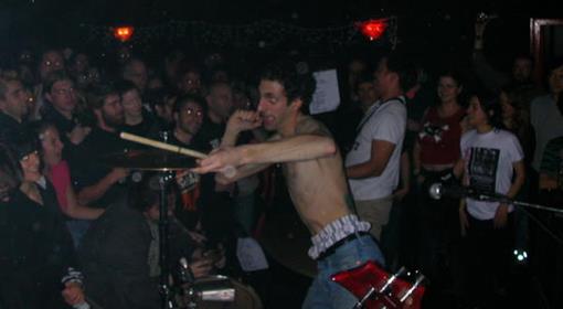 A shirtless drummer performs energetically in front of a lively crowd at a dimly lit indoor concert, raising a drumstick dramatically while people watch closely.