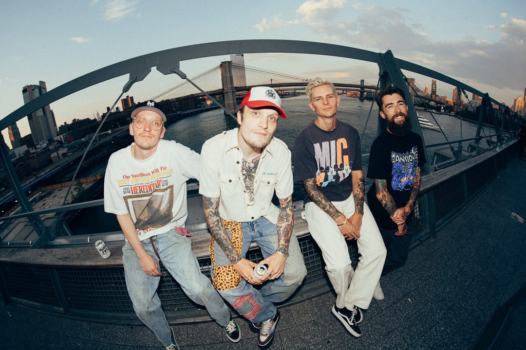 Four young men with tattoos and casual clothes sit on a railing of a bridge at sunset, smiling and relaxed, with a cityscape and river in the background.