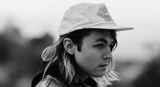 A person with long hair, wearing a baseball cap and a nose ring, looks thoughtfully to the side. The photo is black and white, with a blurred background.