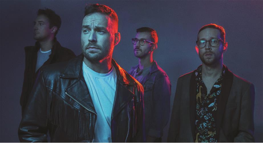 Four men stand together in moody blue and pink lighting. One man in front wears a leather jacket and white shirt, while the other three stand behind him, wearing glasses, jackets, and patterned clothing.