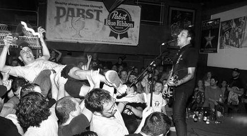 A black-and-white photo of a lively concert crowd-surfing in front of a guitarist and singer on stage, with Pabst Blue Ribbon banners in the background.