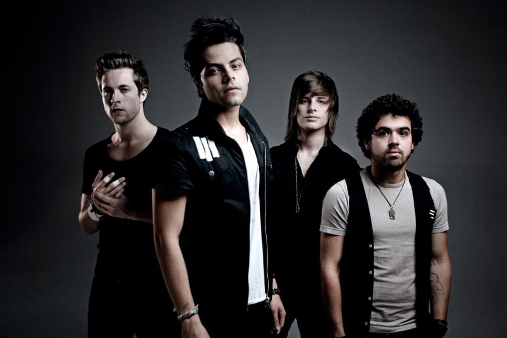 Four young men stand outdoors near water, wearing casual clothing and necklaces. Two have short hair, one wears a beanie, and one has longer, dark hair. The background shows docks and a lakeside structure.
