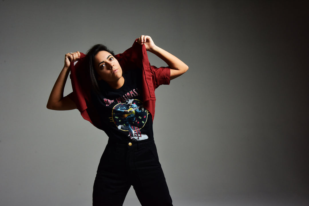A person stands against a plain background, lifting a red jacket over their shoulders. They wear a black graphic t-shirt and high-waisted pants, gazing upward with a confident expression.