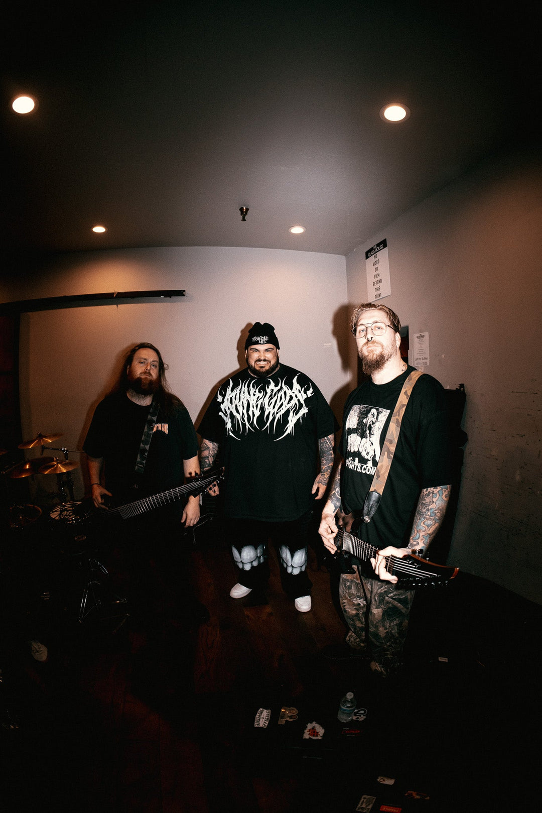 Three men with guitars stand in a dimly lit room, facing the camera. They wear black shirts and have long hair or beards. Musical equipment is visible in the background.