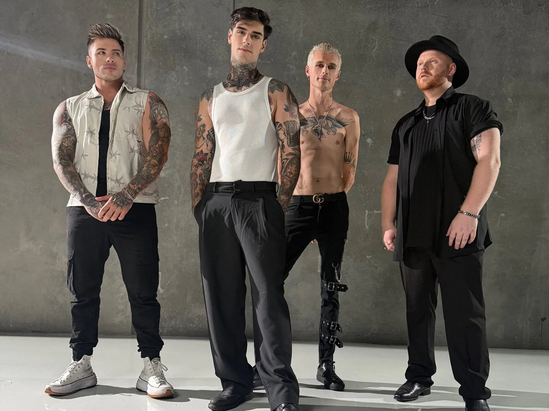 Four men with tattoos stand against a gray wall; one wears a white vest, another a white tank top, the third shirtless with leg accessories, and the fourth in black with a hat.