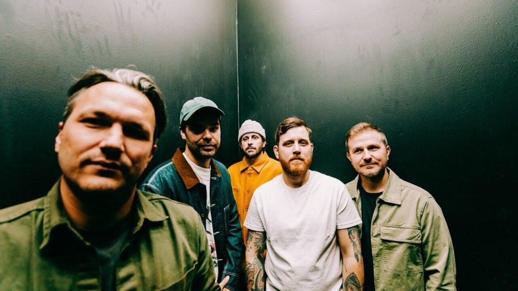 Five men stand close together against a dark, plain wall, looking at the camera. They wear casual jackets, shirts, and hats, with neutral or serious expressions.