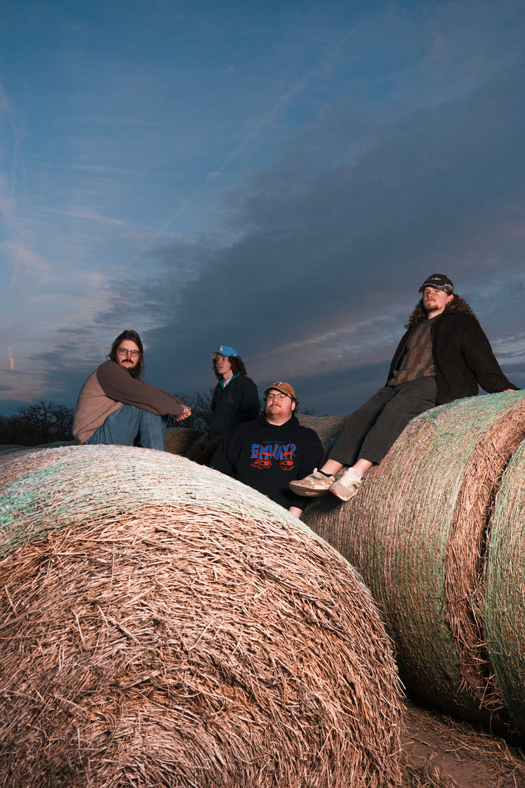 Four people sit on large hay bales outdoors at dusk, with a dramatic cloudy sky behind them. They wear casual clothes and jackets, looking relaxed in the rural setting.