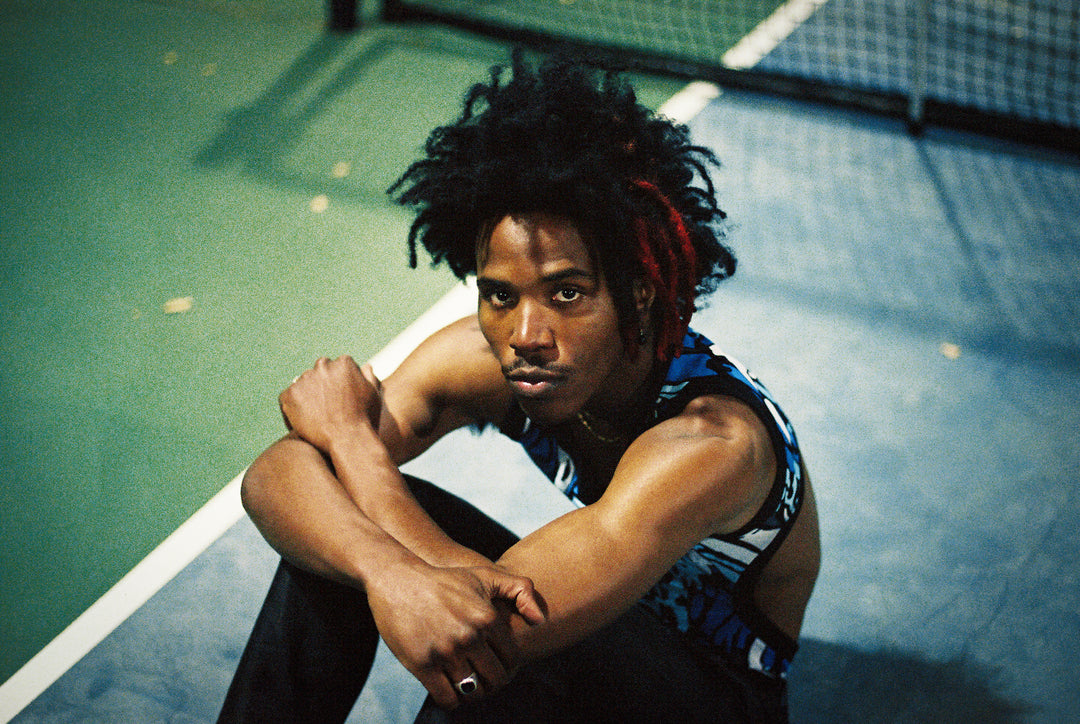 A person with dark, curly hair and a streak of red sits on a tennis court, arms resting on their knees, looking intently at the camera. They wear a sleeveless patterned top and dark pants.