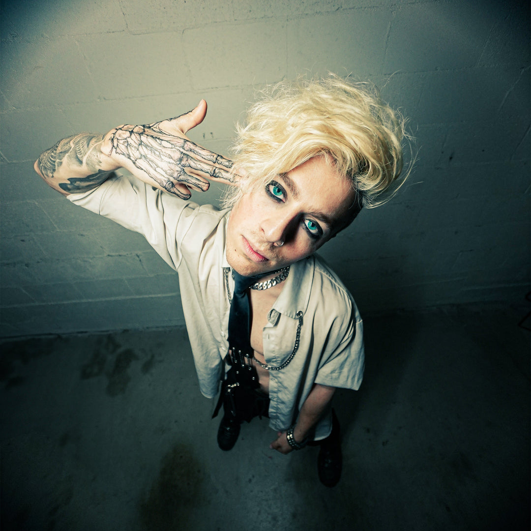 A person with platinum blonde hair, dramatic eyeliner, and tattoos poses indoors, holding their tattooed hand to their head like a gun. The photo is taken from above with a wide-angle lens.