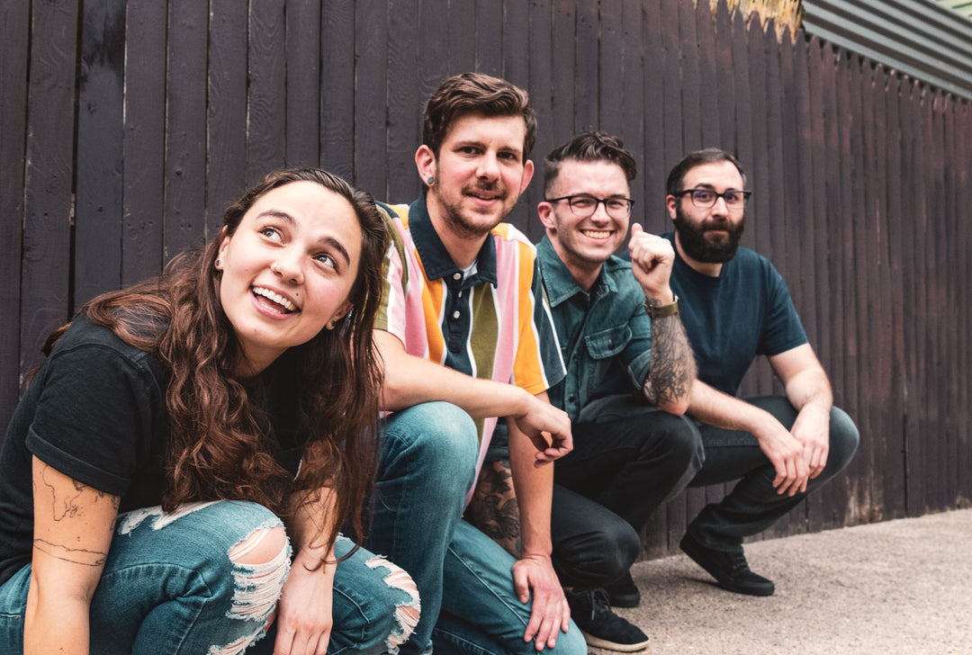 Four young adults, three men and one woman, crouch and smile outdoors in front of a dark wooden fence, appearing happy and relaxed.
