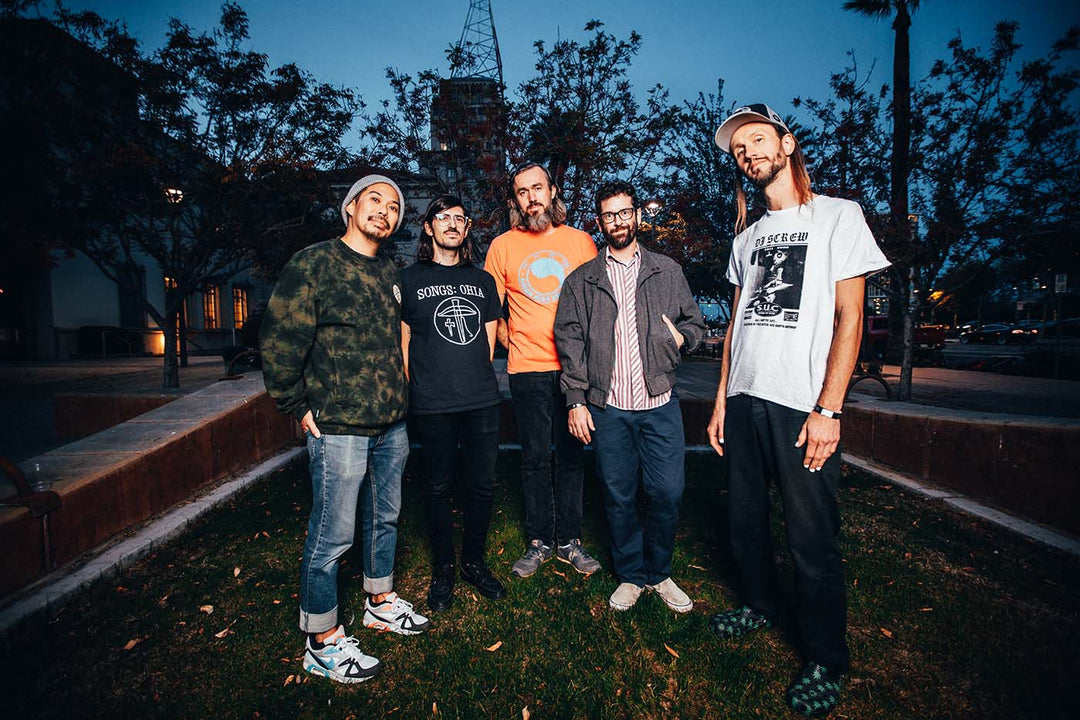 Five men stand together outdoors in a park at dusk, smiling at the camera. Trees and buildings are visible in the background under a darkening sky.