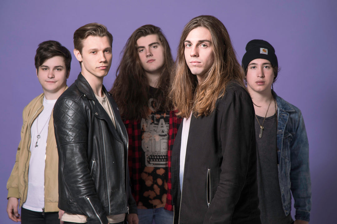 Five young men stand together against a purple background, wearing casual and rocker-style clothes, with varying hairstyles. They look directly at the camera with neutral or serious expressions.