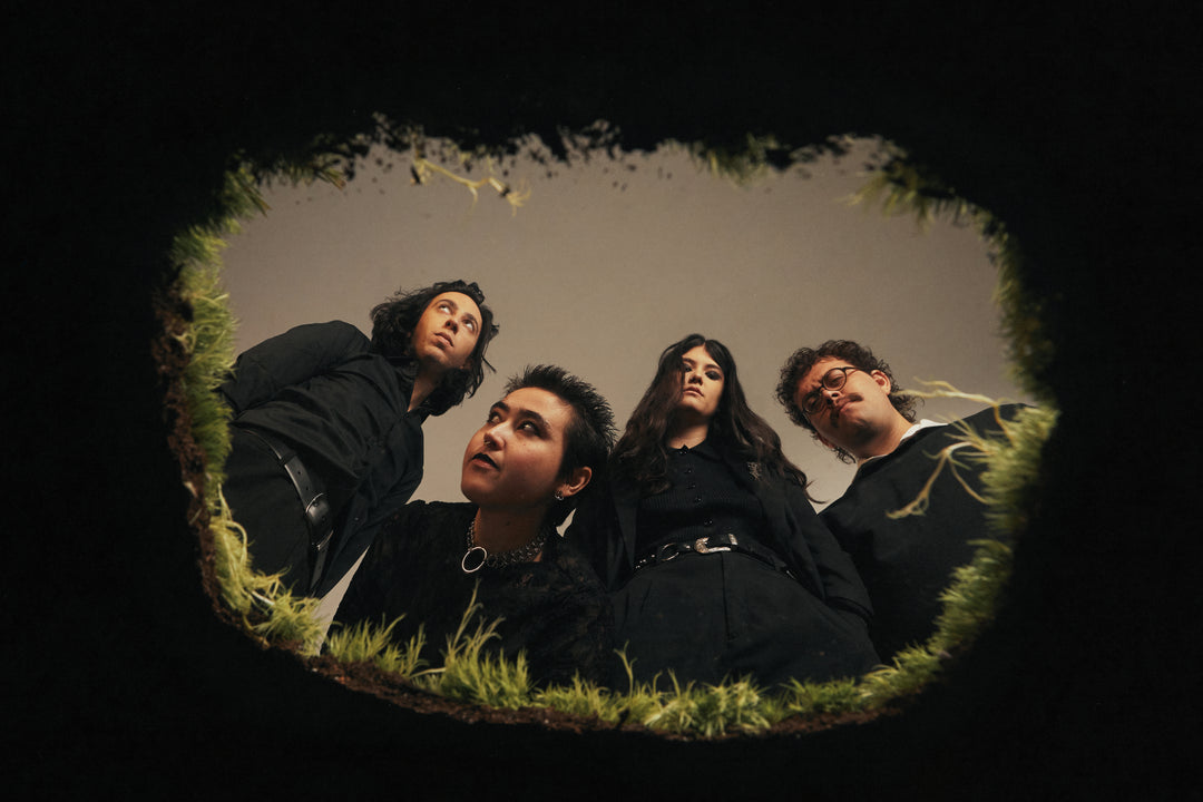 Four people dressed in black stand close together, looking down into a hole in the ground framed by grass and dirt, as if the photo is taken from inside the hole looking up at them.