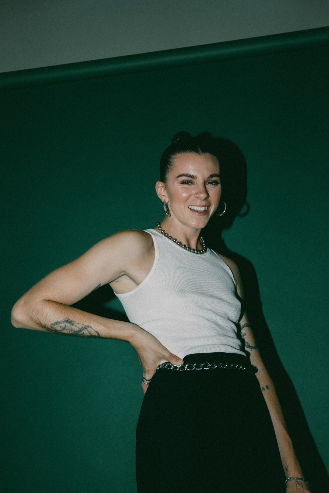 A smiling person with short dark hair, wearing a white tank top, black pants, and chain accessories, stands in front of a dark green background with one hand on their hip.