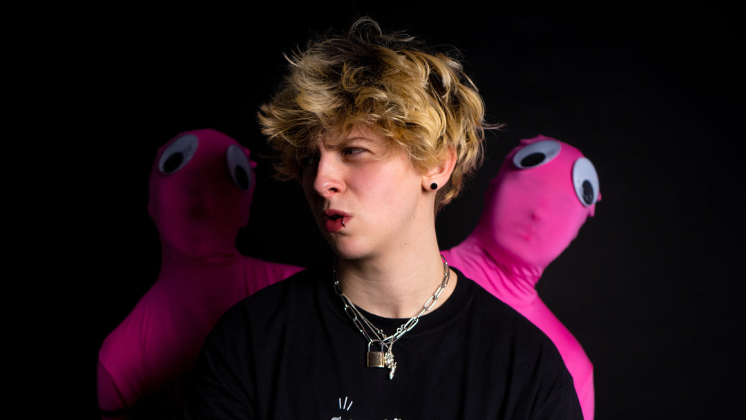 A person with curly blond hair and layered necklaces stands in front of two figures in pink bodysuits with large googly eyes against a black background. The person looks to the side with a surprised expression.