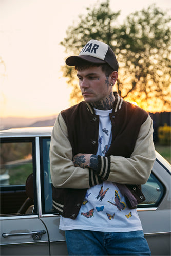 A young man with tattoos, wearing a black and beige varsity jacket, butterfly T-shirt, and cap, stands with crossed arms beside a car at sunset, with trees and a soft sky in the background.