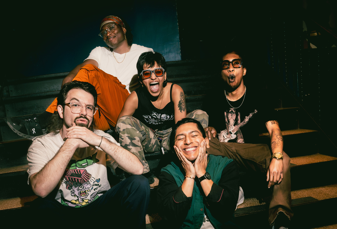 Five people pose joyfully on stairs, smiling and making playful faces. They wear casual clothes, including graphic tees, sunglasses, and colorful pants, in a lively, relaxed atmosphere.