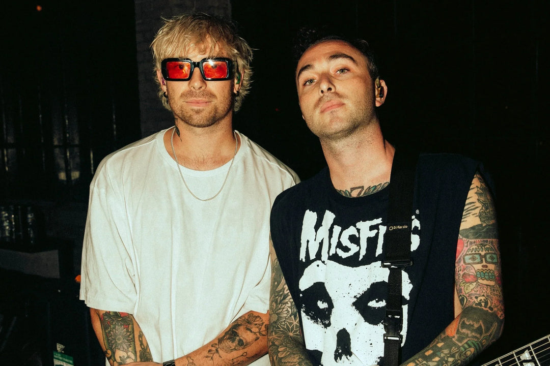 Two men with tattoos stand together indoors. One wears sunglasses and a white shirt, the other wears a sleeveless Misfits shirt. Both look at the camera with serious expressions.