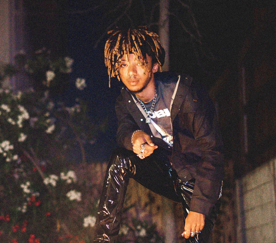 A person with blond dreadlocks, wearing a black jacket and shiny black pants, crouches outdoors at night with plants and flowers blurred in the background.